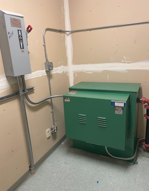 The image shows a small, green electrical generator connected to a wall outlet, with a switch and a warning sign, situated in a plain, white room.AI-generated content may be incorrect.