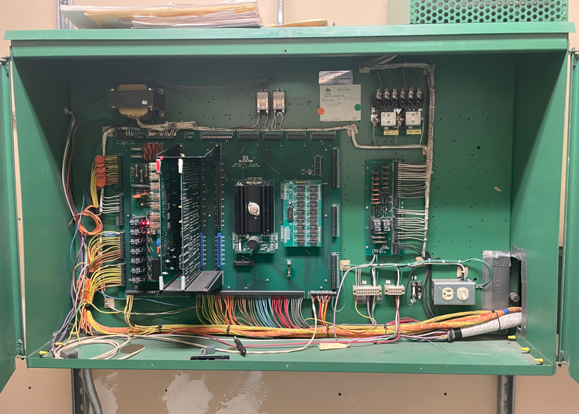 The image shows a complex, green control panel with multiple switches, buttons, and indicator lights, likely part of a control system.AI-generated content may be incorrect.