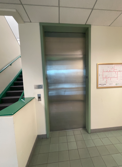 The image depicts a metallic door leading to an elevator, with a stairwell visible in the background and a wall chart to the right.AI-generated content may be incorrect.
